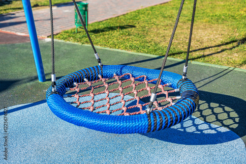 A blue round rope swing with a red net is suspended on a modern playground. The area features a rubberized surface for safety, surrounded by greenery and paths.