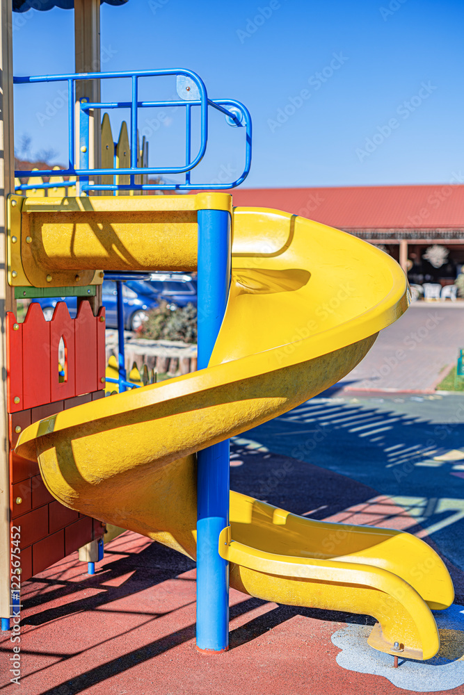 A vibrant yellow spiral slide stands in the playground, surrounded by bright blue metal railings and a colorful play structure.