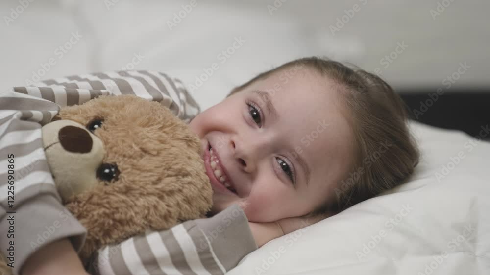 Child smiles happily and hugs plush teddy bear tightly lying on pillow in bedroom. Daughter preschooler wants mother to tell fairytale before falling asleep Child smiles happily and hugs plush teddy bear tightly lying on pillow in bedroom. Daughter preschooler wants mother to tell fairytale before falling asleep