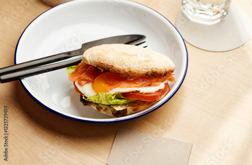 A delicious breakfast sandwich is served on a white plate in a cozy cafe. The sandwich includes a soft bun filled with cooked egg, smoked salmon, lettuce, and tomato photo