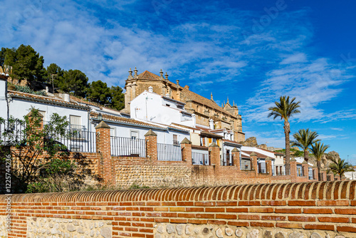 Cityscape with brick wall and wrought iron railings on street on hillside, houses and Royal Collegiate Church of Santa María la Mayor in background, sunny day in Antequera, Spain photo