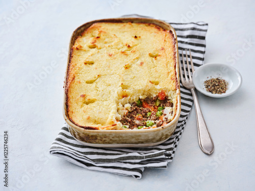 A hearty shepherd's pie is showcased, featuring a fluffy mashed potato topping and a savory meat filling with vegetables. It rests on a striped cloth with seasoning nearby photo