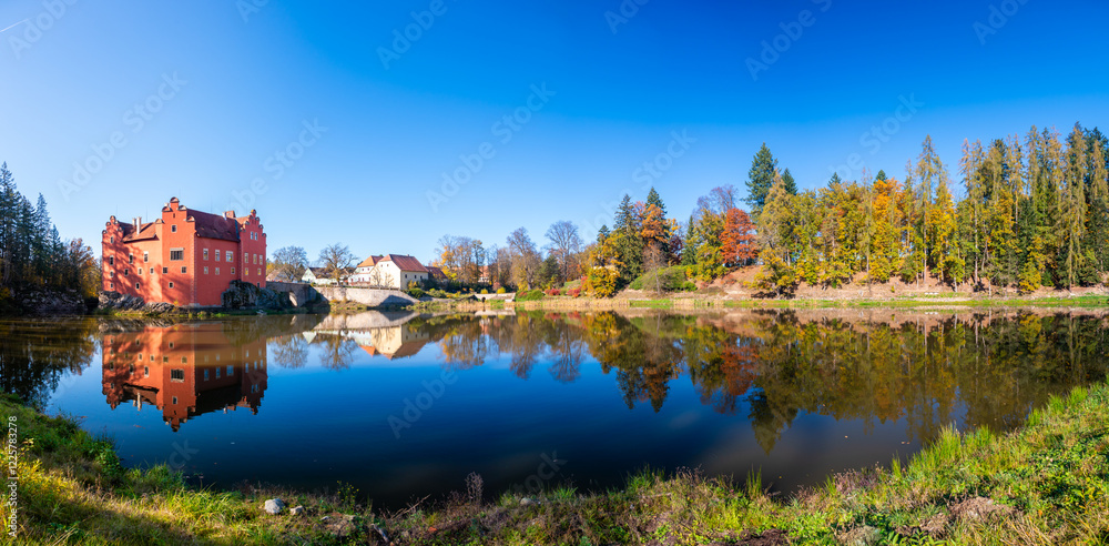 Panoramic view of Cervena Lhota Castle in the Czech Republic, surrounded by a lake reflecting its image. Autumn trees in vibrant colors and sunny weather create a picturesque, serene scene.