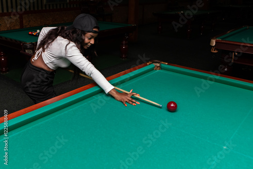 Young black woman wearing a baseball cap is playing billiards, aiming for the red ball with her cue stick on a green billiard table in a pool hall, demonstrating concentration and skill photo