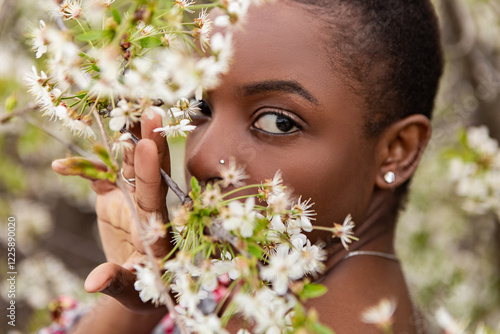 Beautiful young model with short hair and nose piercing gently touching and smelling delicate white cherry blossoms on a flowering tree branch in an orchard in springtime. Meditation, mental treatment