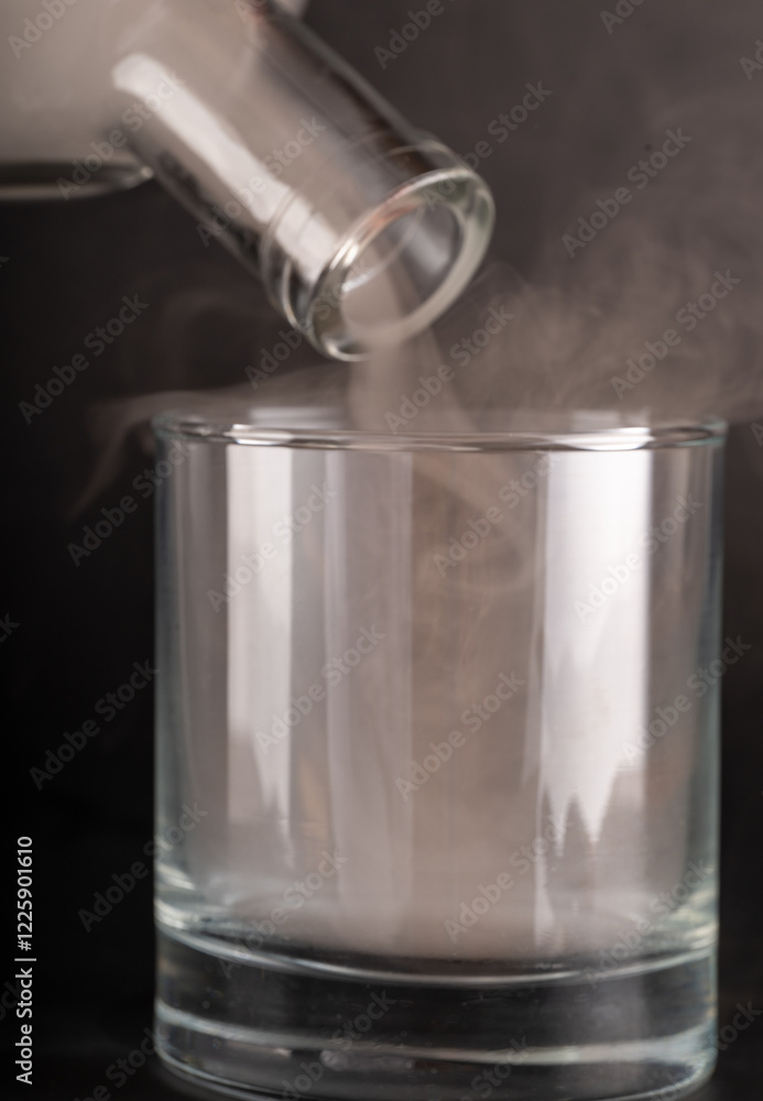 A close-up shot of a clear glass tumbler being filled with thick, white vapor. The vapor pours from a tilted glass bottle, overflowing the glass. The background is a soft-focus, dark gray. A close-up shot of a clear glass tumbler being filled with thick, white vapor. The vapor pours from a tilted glass bottle, overflowing the glass. The background is a soft-focus, dark gray.