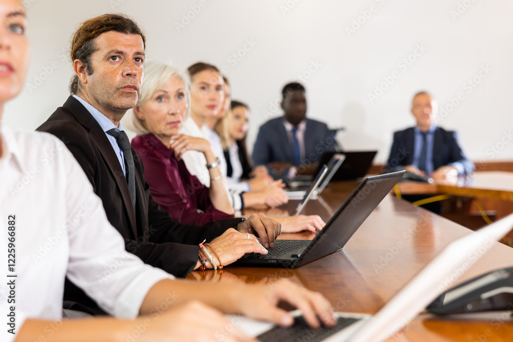 Focused mature white business man in formal suit sitting with colleagues in conference room and absorbedly listening to speaker's presentation during corporate group meeting Focused mature white business man in formal suit sitting with colleagues in conference room and absorbedly listening to speaker's presentation during corporate group meeting