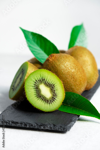 Ripe kiwi fruits in sunlight. Heap of whole kiwi fruits with green leaves and fresh cut kiwi on slate board. Healthy organic fruits full of vitamins and antioxidants.
