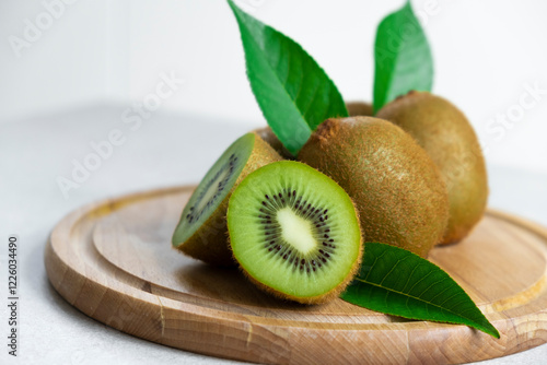 Ripe kiwi fruits in sunlight. Heap of whole kiwi fruits with green leaves and fresh cut kiwi on wooden cutting board. Healthy organic fruits full of vitamins and antioxidants.