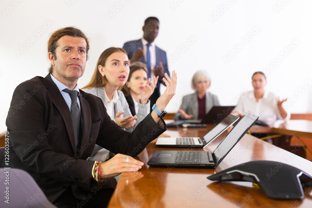 Nervous man in suit sitting at desk with group of coleagues during meeting. Nervous man in suit sitting at desk with group of coleagues during meeting.