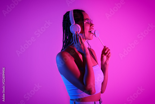 Young African-American woman with lollipop listening to music on neon background photo