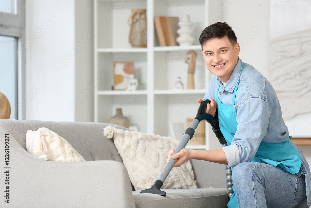 Obraz premium Young man in apron vacuum cleaning sofa at home