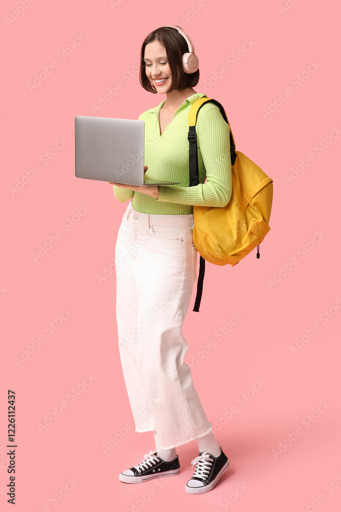 Fototapeta premium Beautiful young woman in headphones working with modern laptop on pink background