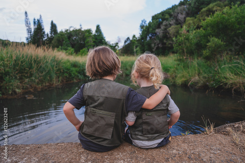Two siblings in adventuring clothing hugging each other in front of water