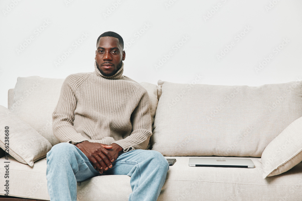 African man in a cozy turtleneck sweater sitting on a light colored sofa, reflecting calmness and introspection in a modern interior setting African man in a cozy turtleneck sweater sitting on a light colored sofa, reflecting calmness and introspection in a modern interior setting