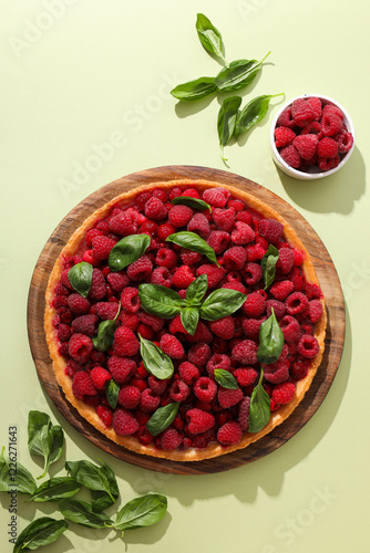 Berry pie on wooden plate, raspberries in bowl and basil leaves on light green background, top view photo