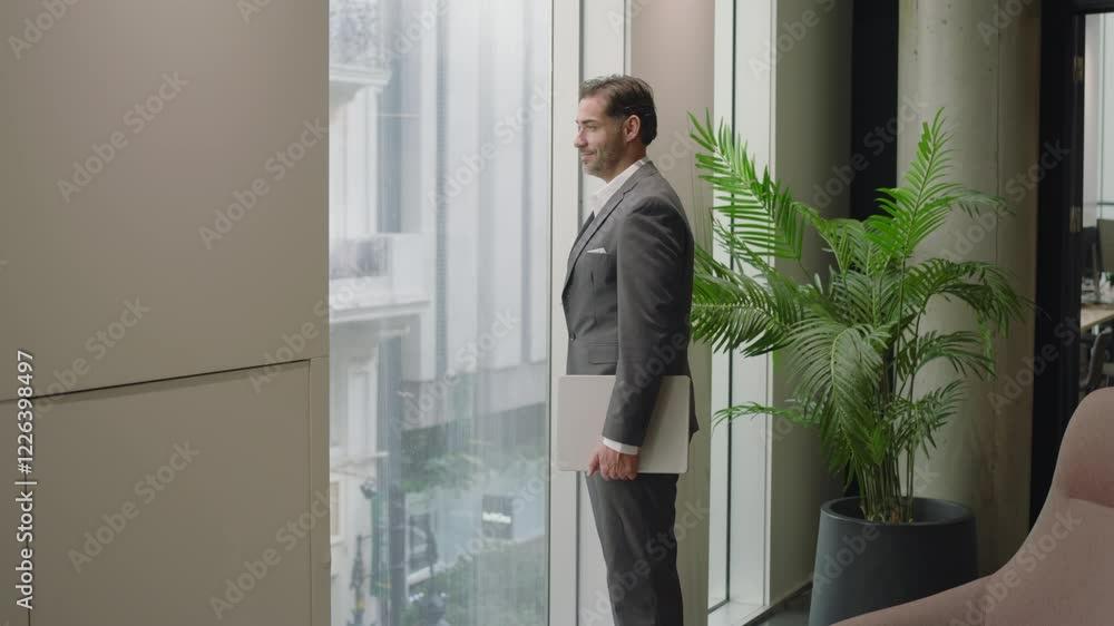 Confident businessman in tailored suit stands with laptop by floor to ceiling windows, observing the city below, while a lush green plant adds a vibrant touch to the sophisticated office ambiance Confident businessman in tailored suit stands with laptop by floor to ceiling windows, observing the city below, while a lush green plant adds a vibrant touch to the sophisticated office ambiance
