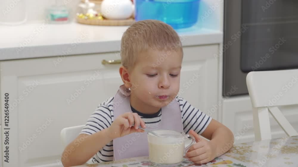 Child eating yogurt with spoon from glass cup at dinner table at home. Happy little boy eating ryazhenka with appetite. Satisfied kid smiling. Child eating yogurt with spoon from glass cup at dinner table at home. Happy little boy eating ryazhenka with appetite. Satisfied kid smiling.