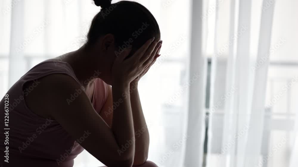 Sad woman covers face with hands sitting against window Sad woman covers face with hands sitting against window
