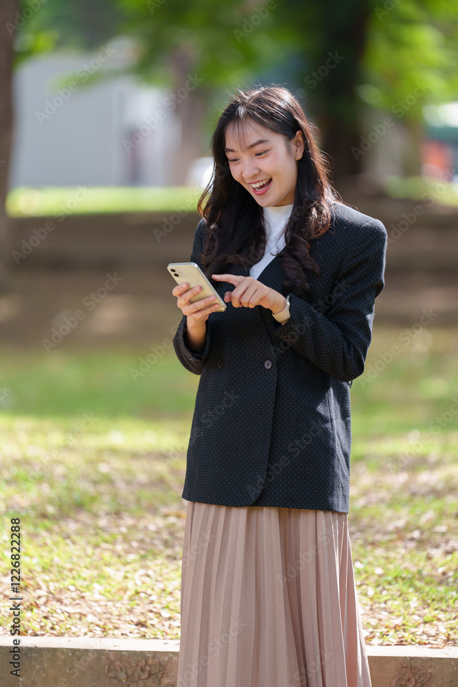 Young asian businesswoman wearing a stylish blazer and pleated skirt, smiling while using her smartphone in a vibrant green park on a sunny day, enjoying the outdoors and modern connectivity Young asian businesswoman wearing a stylish blazer and pleated skirt, smiling while using her smartphone in a vibrant green park on a sunny day, enjoying the outdoors and modern connectivity