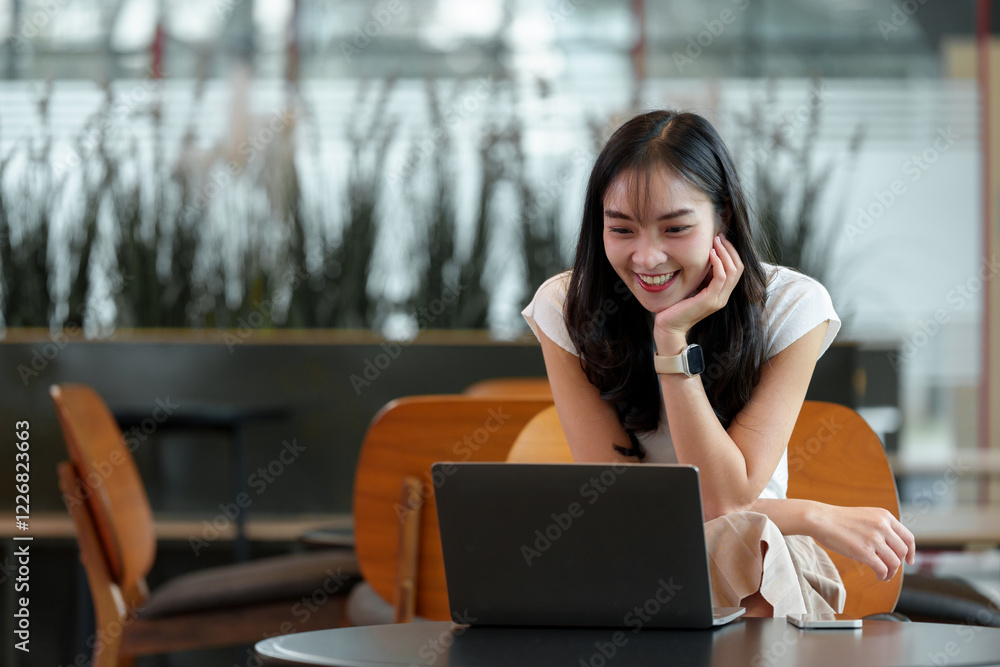 Naklejka premium Smiling young woman using a laptop at a cafe, showcasing remote work or online learning. Modern setting with natural light and comfortable seating, highlighting digital connectivity