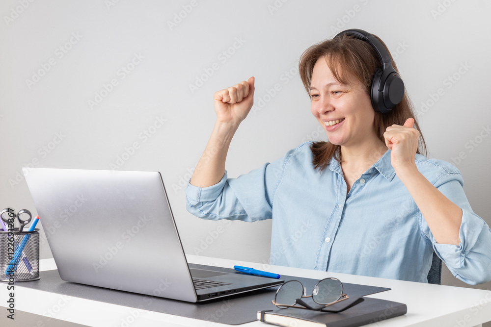 Success, victory or meeting deadline concept. Smiling excited caucasian woman freelancer with headphones behind a laptop is holding her hands up in the air. Success concept Success, victory or meeting deadline concept. Smiling excited caucasian woman freelancer with headphones behind a laptop is holding her hands up in the air. Success concept