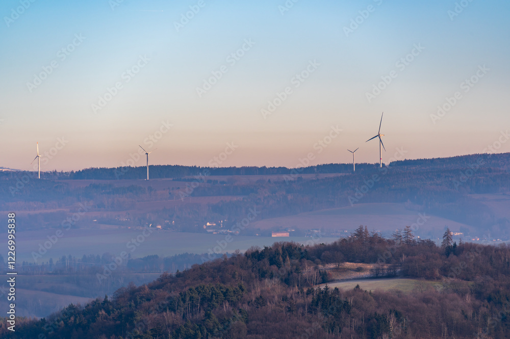 Wind turbines stand tall on rolling hills during a stunning sunset. The sky transitions from deep blue to vibrant shades of purple and red, creating a dramatic and peaceful scene