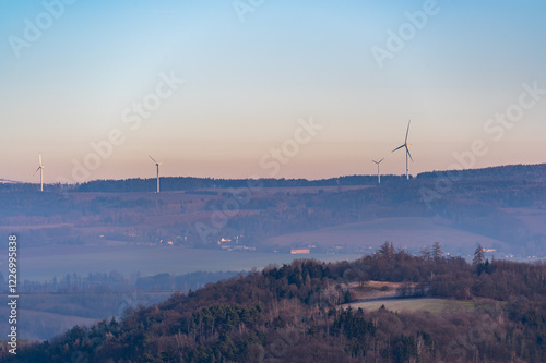 Obraz na płótnie Wind turbines stand tall on rolling hills during a stunning sunset. The sky transitions from deep blue to vibrant shades of purple and red, creating a dramatic and peaceful scene w salonie
