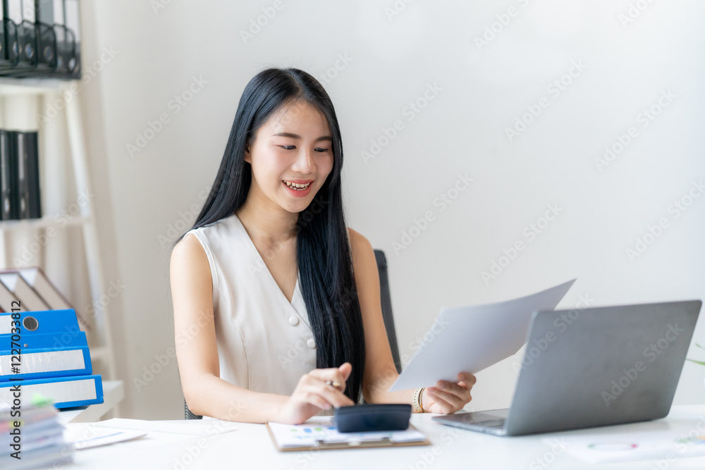 Focused and Efficient: A young businesswoman smiles confidently as she works on her laptop and examines documents, showcasing dedication and professionalism in her modern workspace. Focused and Efficient: A young businesswoman smiles confidently as she works on her laptop and examines documents, showcasing dedication and professionalism in her modern workspace.