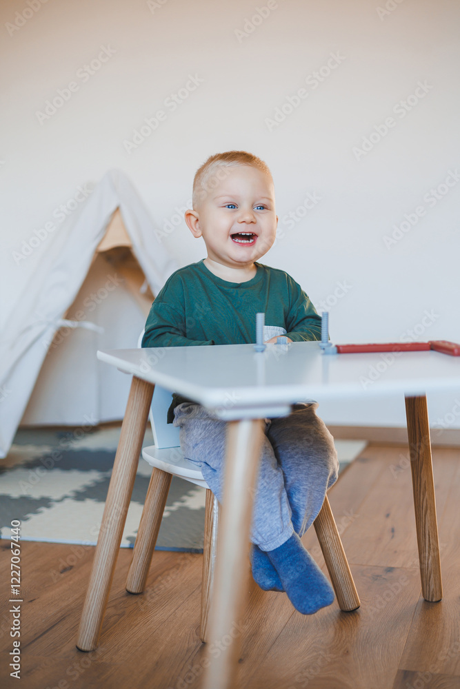 Blond boy toddler playing with toys in children's room close-up and copy space. Child sitting on a stool at a table. Furniture for children Blond boy toddler playing with toys in children's room close-up and copy space. Child sitting on a stool at a table. Furniture for children