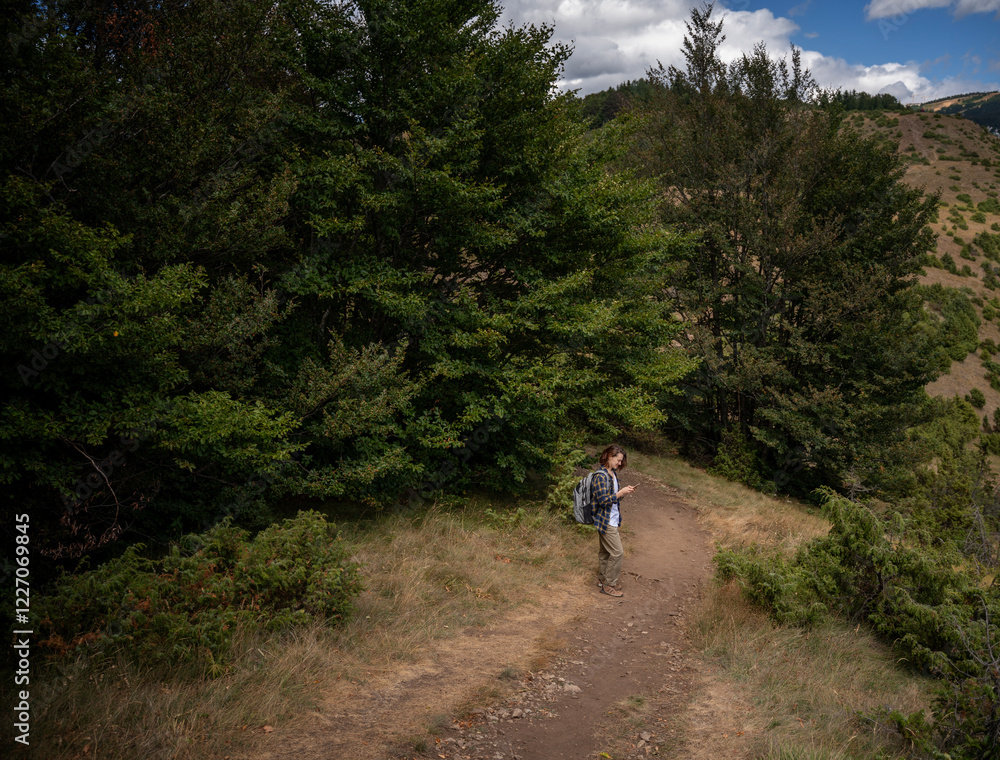Young woman traveler trekking on forest mountain trail in summer using smartphone Young woman traveler trekking on forest mountain trail in summer using smartphone