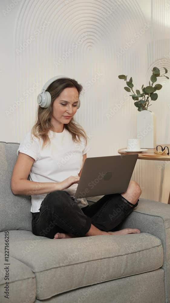 Vertical video, cheerful woman sits on the couch at home, listening to music on headphones, using a laptop. Vertical video, cheerful woman sits on the couch at home, listening to music on headphones, using a laptop.