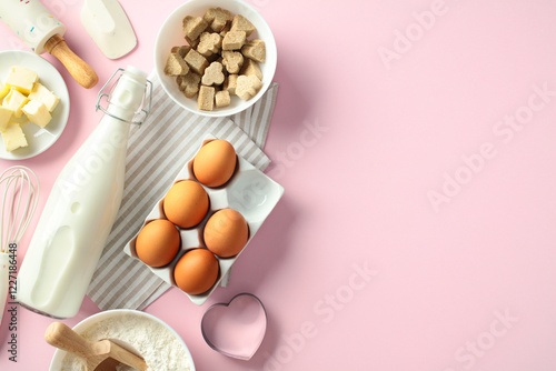 Flat lay baking ingredients on a pink background: milk bottle, eggs, butter, flour, sugar cubes, whisk, spatula, and rolling pin photo