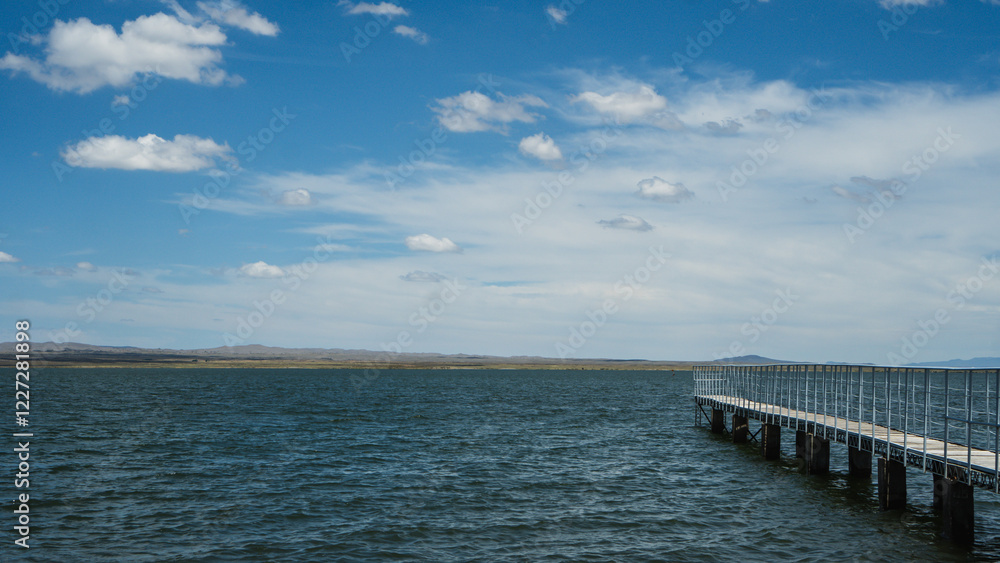 Fototapeta premium A long wooden dock extends into a calm lake on a sunny day. The water is a deep blue-green color and there are ripples on the surface. The sky is clear with a few white clouds.