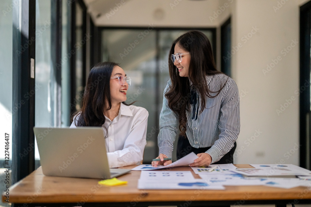 Two women are sitting at a desk with a laptop and papers in front of them Two women are sitting at a desk with a laptop and papers in front of them