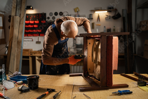Bild auf Leinwand Elderly male furniture repair restorer specialist in uniform uses electric cordless screwdriver, making upholstery vintage chair restoring armchair in workshop