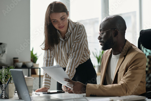 Young businesswoman bending over workplace and looking at financial paper document held by male colleague photo