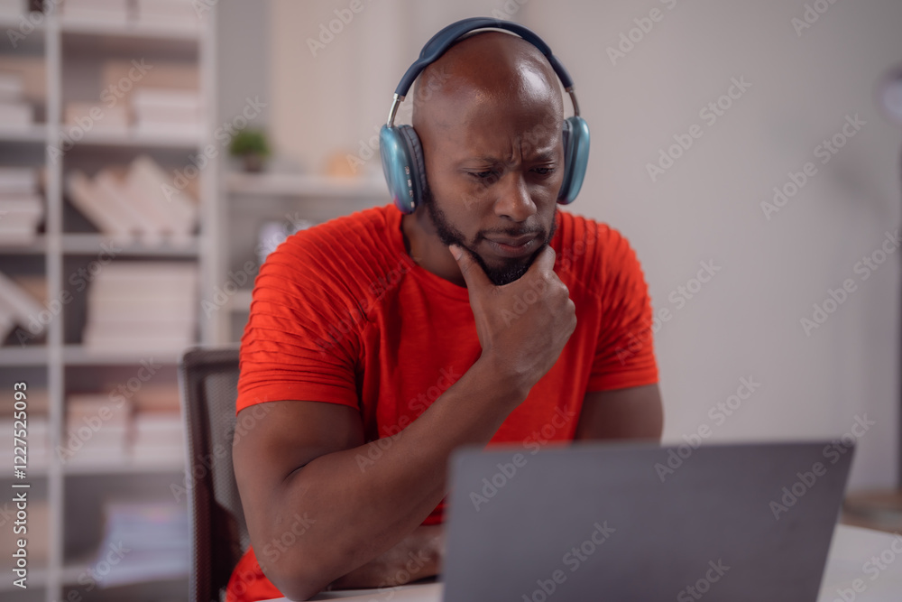 Focused black man wearing headphones is working from home, using a laptop and thoughtfully resting his hand on his chin while contemplating ideas and strategies for his project Focused black man wearing headphones is working from home, using a laptop and thoughtfully resting his hand on his chin while contemplating ideas and strategies for his project