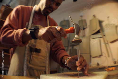 Man focused intently while hammering nail on woodworking project in cozy workshop. Wearing apron and using proper tools. Various woodworking tools visible in background photo