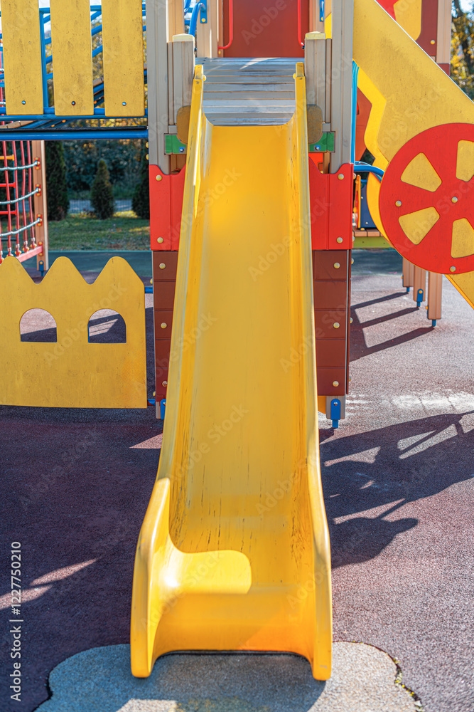 A vibrant yellow spiral slide stands in the playground, surrounded by bright blue metal railings and a colorful play structure.
