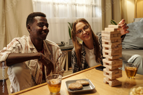 African American guy looking at his girlfriend putting block on top of wooden tower during tabletop game photo