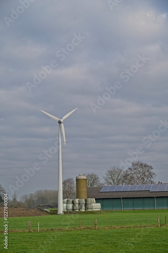 Windmill and solar panels on the roof of a stable. There is a silo at the stable and there are plasticized round bales of hay. photo