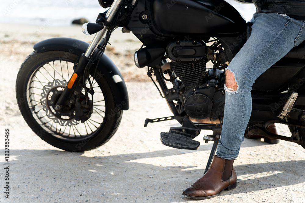 Biker wearing ripped jeans and chelsea boots sitting on a motorcycle on the beach Biker wearing ripped jeans and chelsea boots sitting on a motorcycle on the beach