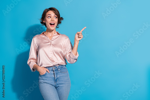 Smiling young woman in a satin pink blouse pointing with excitement against a blue background