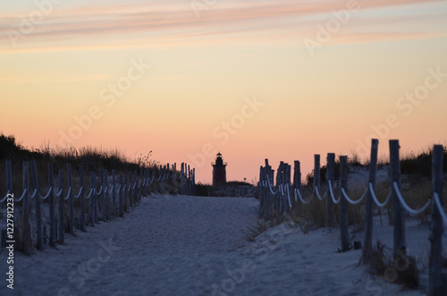 Fototapeta Naklejka Na Ścianę i Meble -  Sandy beach path with rope rails leading to lighthouse silhouette sunset landscape