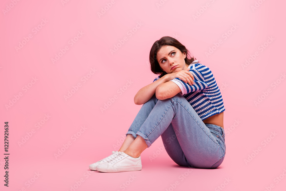 Reflective young woman in stylish striped top sitting on pink background, conveying relaxed and thoughtful vibe Reflective young woman in stylish striped top sitting on pink background, conveying relaxed and thoughtful vibe