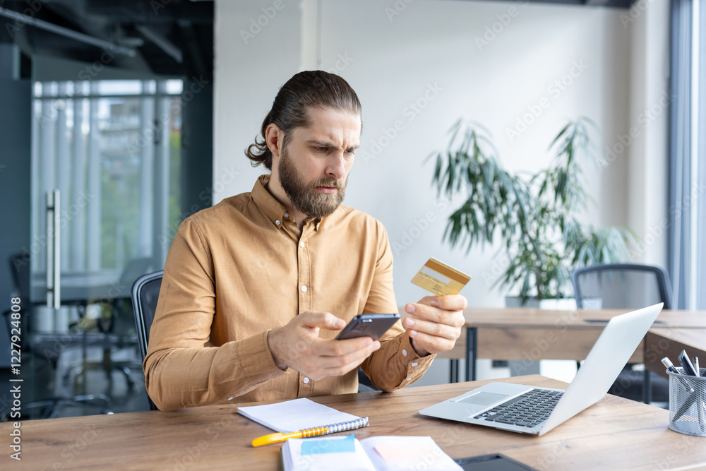 Focused man sitting at a desk in an office environment interacting with a smartphone and a credit card, implying online payment, banking, or identity verification. Focused man sitting at a desk in an office environment interacting with a smartphone and a credit card, implying online payment, banking, or identity verification.