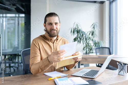 A smiling businessman sitting at a desk in an office, opening an envelope while using a laptop. The scene depicts professionalism, office work, and a friendly workplace atmosphere. photo