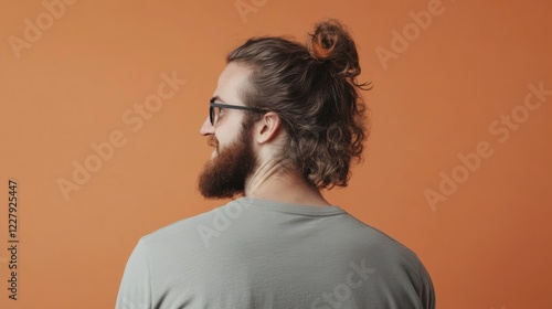 A modern portrait of a young man with curly hair and a beard, wearing a casual shirt and glasses, smiling while standing against an orange background. photo