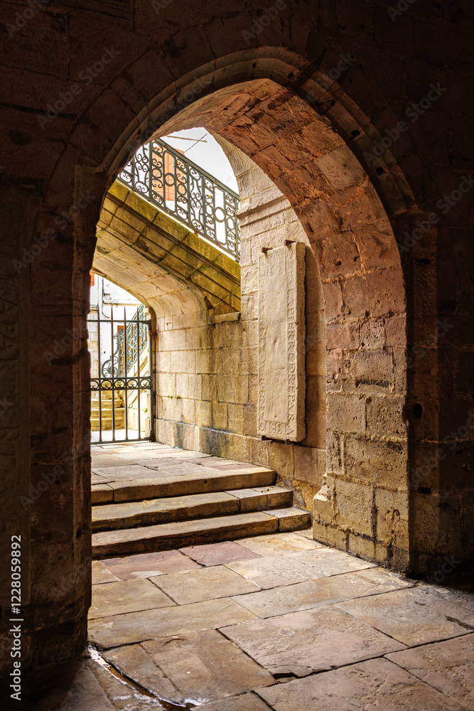 Fototapeta premium Arched stone passage leading to the entrance of Saint-Jean-Baptiste Church in Saint-Jean-de-Luz, France, featuring a historical stone slab and an ornate staircase with wrought iron railing.